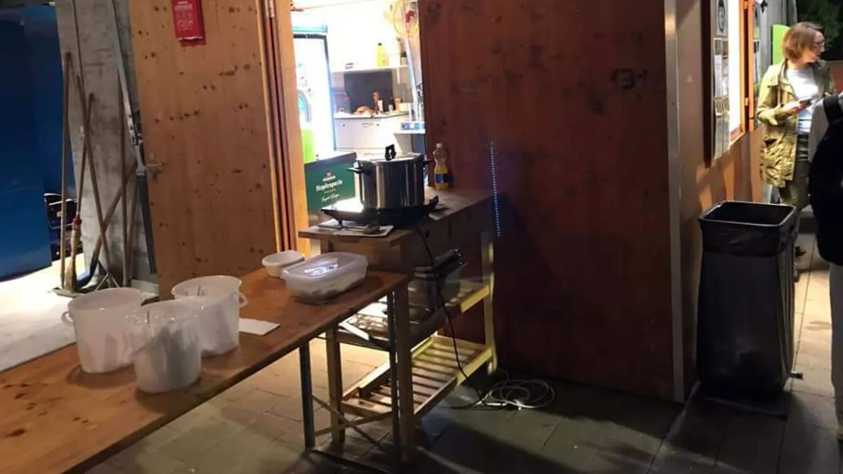 Ein Holztisch steht vor einem Materialraum. Im Hintergrund ist eine Person zu sehen, die aus einem Fenster bedient wird.